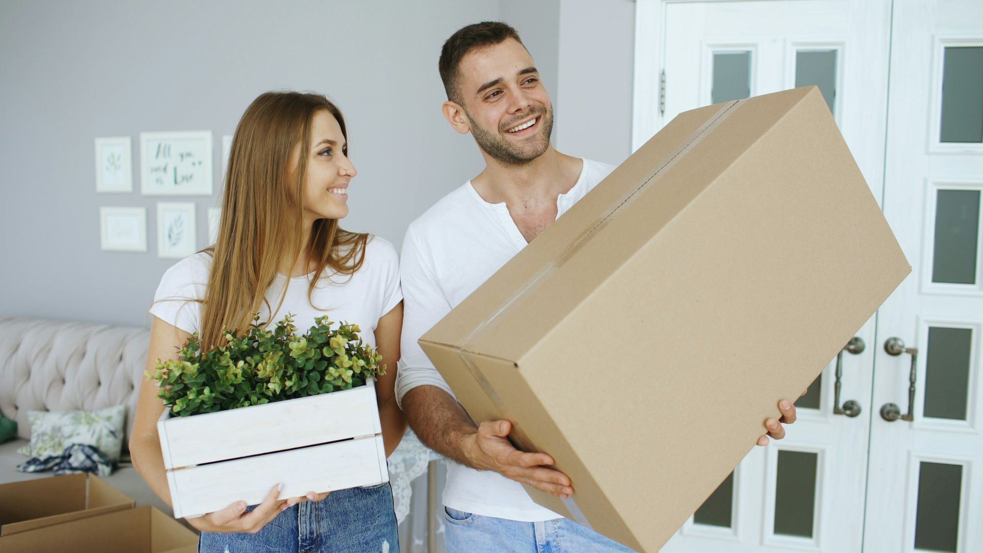 Pareja sonriente llevando una caja de mudanza y una planta a su nueva casa